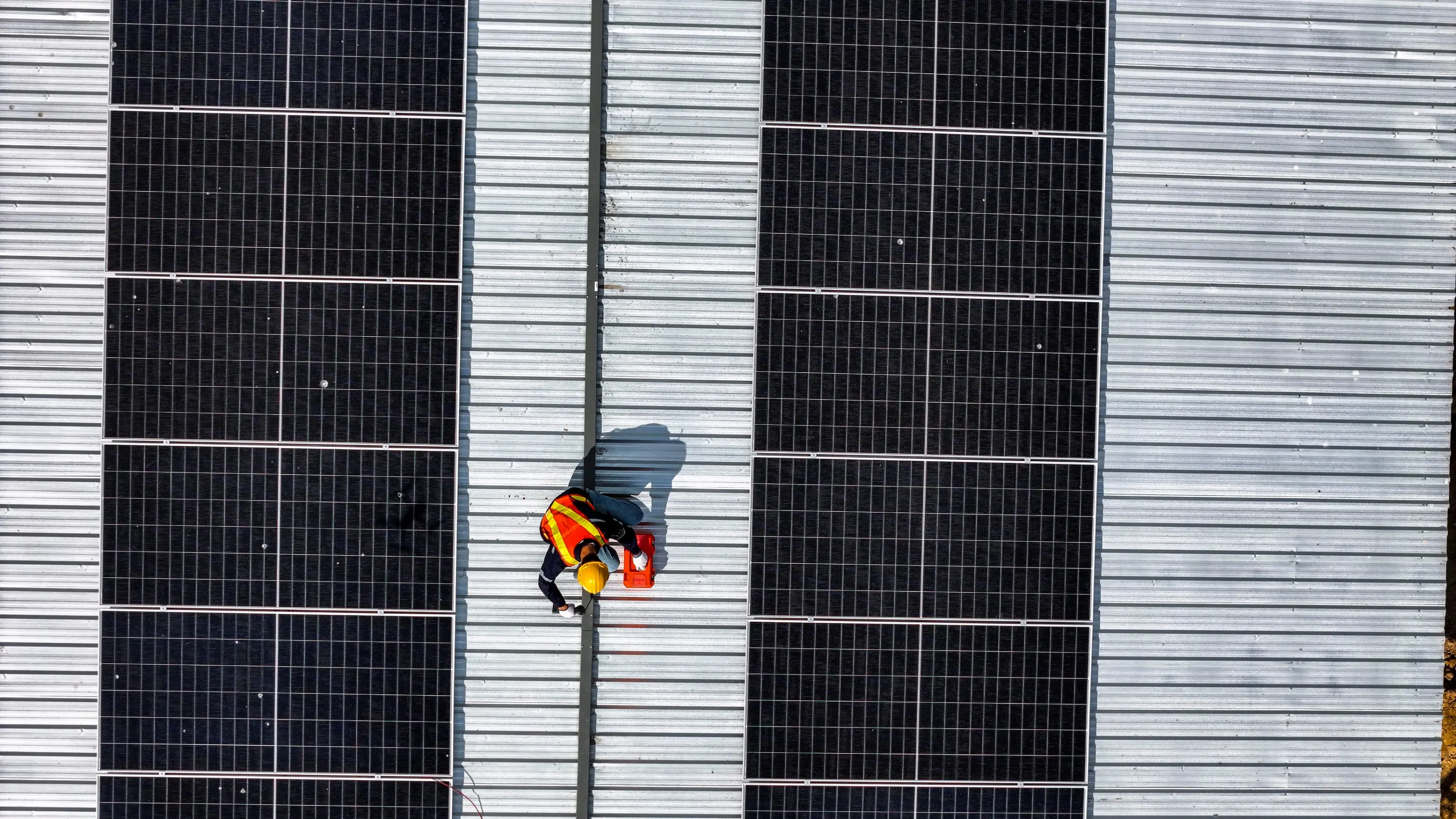 Roofer inspecting a commercial metal roof between rows of solar panels, seen from above